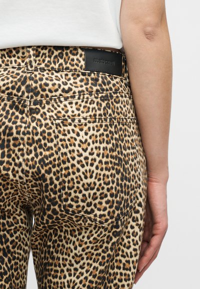 Κοντινό πλάνο ατόμου που φοράει παντελόνι με λεοπάρ σχέδιο, πίσω τσέπη και μαύρη ετικέτα μάρκας, και λευκό πουκάμισο μερικώς ορατό.