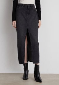 Falda maxi de mezclilla negra con una abertura frontal, cintura alta y detalles de costura sutiles. Combinada con botas de tobillo negras sobre un fondo neutro.