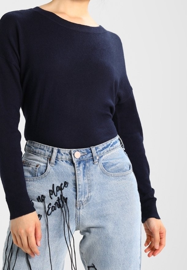 Personne portant un haut à manches longues bleu marine et un jean taille haute bleu clair avec une broderie noire et des fils pendants.