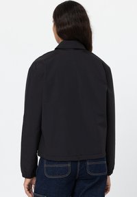 Chaqueta negra con cuello, mangas largas, puños elásticos y un ajuste corto, hecha de una tela suave, sin patrones visibles.