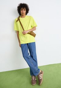 Camiseta amarilla neón de gran tamaño, pantalones vaqueros anchos azules, zapatillas marrones con rayas blancas y un bolso cruzado marrón sobre un fondo liso.