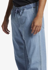 Svjetloplave traperice s elastičnim strukom, bočnim džepovima i vezicom. Tkanina ima glatku teksturu i opušteni kroj.