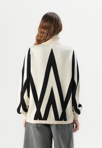 Femme portant un pull à col roulé crème avec un motif zigzag angulaire noir audacieux et un pantalon gris ample, vue de dos.