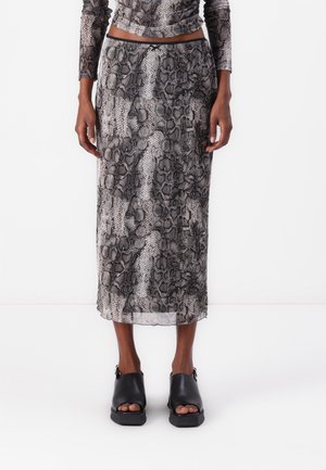 Vrouw draagt een grijze en zwarte rok met slangenprint, hoog in de taille en tot boven de enkels, gecombineerd met zwarte open-toe sandalen met blokhak.
