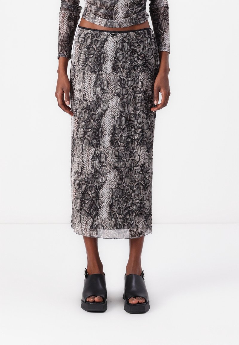 Femme portant une jupe à motifs serpent gris et noir, taille haute, longueur cheville, avec des sandales à talons blocs noirs et bout ouvert.