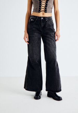 Kobieta nosi czarne dżinsy z szerokimi nogawkami, czarne botki oraz krótki, bezrękawnik z ozdobnym wiązaniem z przodu, stoi na tle jednolitego tła.