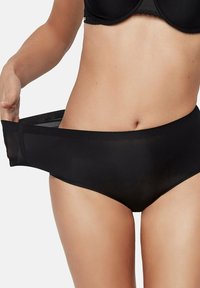 Femme portant des sous-vêtements noirs, tirant la taille pour montrer l'élasticité et l'ajustement du tissu autour des hanches.