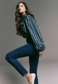 Dlouhý rukáv kostkovaná košile v modré a zelené barvě, s volány; kombinována s tmavě modrými džíny a bílými botami; modelka pózující s jednou nohou zdviženou.