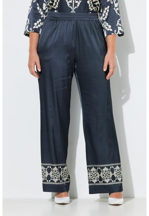 Donna che indossa pantaloni larghi blu navy con stampa di bordo floreale bianco e tacchi bianchi, in piedi contro un muro semplice.