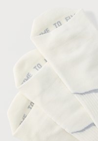 Τρία ζευγάρια λευκές αστραγάλες κάλτσες με γκρι κείμενο και ρίγες στοιβαγμένα σε λευκό φόντο.