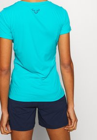 Camiseta deportiva turquesa con cuello redondo, mangas cortas y un pequeño logo en la parte posterior. Combinada con pantalones cortos deportivos azul marino.