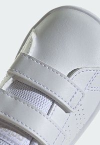 Białe buty sportowe z gładką syntetyczną cholewką, dwoma zapięciami na rzepy, siateczkowym wykończeniem i perforowanymi detalami w okolicy palców.