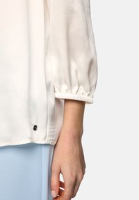 Close-up of a hand beside a white satin blouse with gathered cuff and light blue pants against a plain background.