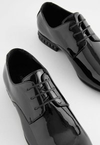 Chaussures habillées en cuir verni noir avec un design classique à lacets, texture lisse et détails subtils sur la semelle.