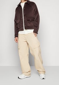 Brun kordfläskjacka med två framfickor, tillsammans med beige cargobyxor och vita sneakers, visad mot en neutral grå bakgrund.