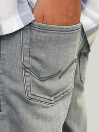 Pantalon en denim gris avec des coutures de poche visibles, une coupe décontractée et un design distinctif des poches arrière. Main placée dans la poche, texture visible.
