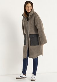 Brauner Kunstpelzmantel mit hohem Kragen, Frontreißverschluss und zwei schwarzen Stofftaschen. Getragen über dunklen Jeans und weißen Sneakers mit schwarzen Streifen.