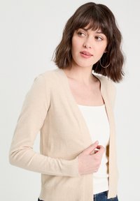 Cardigan en tricot beige avec un design en V, des manches longues, des poignets côtelés et un ourlet droit. Porté par-dessus un haut blanc.