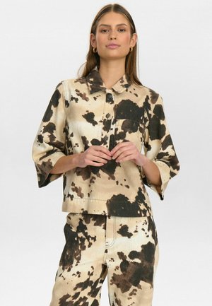 Chemise à col, à manches courtes en tissu léger beige avec un motif de vache brun et noir. Comprend une poche avant et une fermeture à boutons.