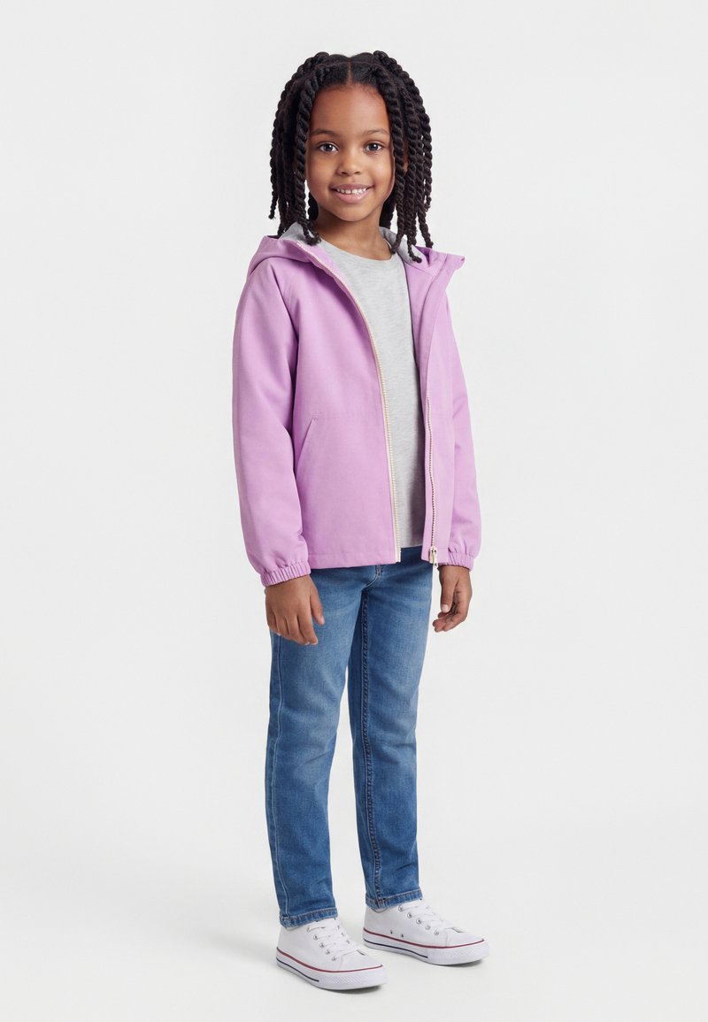 Νεαρή κοπέλα με πλεξούδες, φορώντας ένα ανοιχτό μωβ μπουφάν με κουκούλα, γκρι μπλούζα, μπλε τζιν και λευκά αθλητικά παπούτσια, σε όρθια στάση και χαμογελαστή.