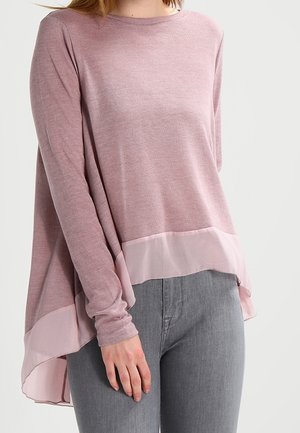Vrouw draagt een mauve trui met lange mouwen en een doorschijnende roze zoom, gecombineerd met lichtgrijze strakke jeans, staand tegen een effen achtergrond.