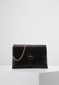 Sac à main en cuir noir avec une finition texturée, une bandoulière en chaîne dorée et un fermoir décoratif proéminent au design en maillons de chaîne.