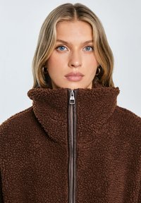 Braune Zip-Jacke aus strukturiertem Fleece-Material, ausgestattet mit hohem Kragen und kontrastierenden Reißverschlussdetails.