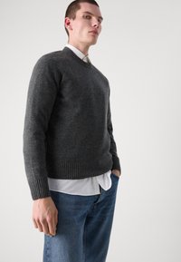 Maglione grigio a maglia con polsini e orlo a coste, indossato sopra una camicia bianca con colletto, abbinato a jeans in denim blu. Design semplice e tessurizzato.