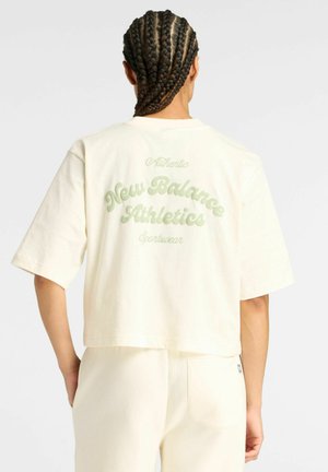 Personne avec des cheveux tressés portant une chemise à manches courtes crème et un pantalon, le dos de la chemise indique "New Balance Athletics Sportswear" en texte vert clair.
