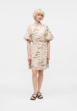 Γυναίκα με κοντά σγουρά ξανθά μαλλιά που φοράει κοντό φόρεμα με ρίγες τίγρης σε μπεζ και καφέ χρώμα, με φουσκωτά μανίκια, μαύρα σανδάλια και χρυσή αλυσίδα στο λαιμό.