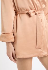 Jasnoróżowy satynowy szlafrok z paskiem w talii, wyposażony w miękkie, pluszowe mankiety oraz gładką fakturę, podkreślający smukły, opływowy design.