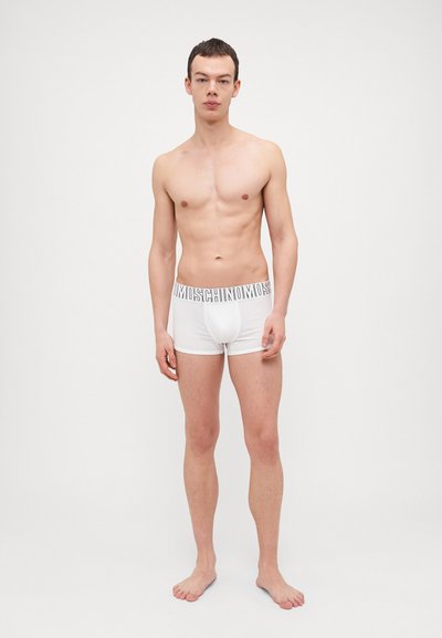 White cotton boxer briefs with a logo waistband. Fitted design, smooth texture, and mid-thigh length. Model stands barefoot against a plain background.