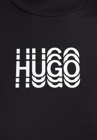 Czarna tkanina z białym stylizowanym napisem "HUGO", obejmującym warstwowe pionowe i krzywe linie, tworzące efekt lustra i cienia.