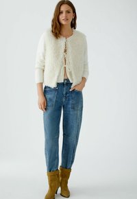 Witte, fluffy vest over een lichte top, gecombineerd met blauwe denimjeans en geelbruine enkellaarsjes. Het vest heeft een ronde halslijn en strikken.