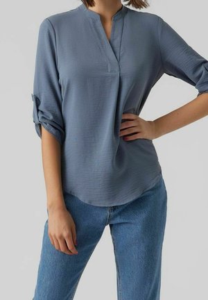 Frau trägt eine lockere, stahlblaue Bluse mit hochgekrempelten Ärmeln und blaue Jeans, steht mit einer Hand in der Hüfte vor einem schlichten Hintergrund.