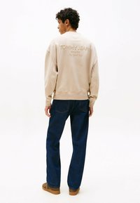 Beige Sweatshirt mit "TOMMY JEANS" und "New York City" auf dem Rücken gestickt, kombiniert mit dunklen blauen Jeans und braunen Schuhen.