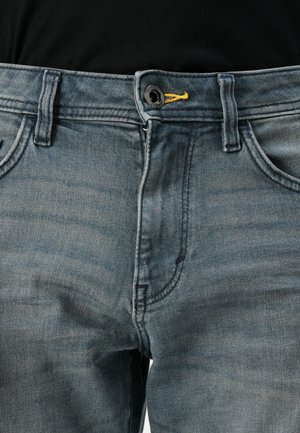 Blugi din denim albastru estompat, cu închidere cu buton, detaliu galben la talie și design clasic cu cinci buzunare. Textura pare moale și purtată.