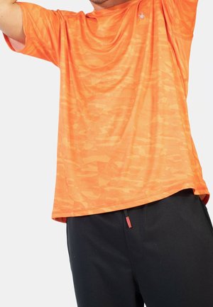 Mand iført orange, mønstret kortærmet skjorte og sorte bukser, løfter armene over hovedet mod en hvid baggrund.