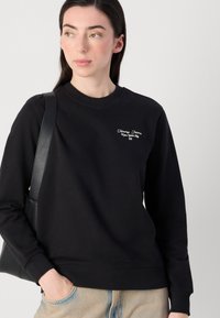 En ung kvinne med langt, mørkt hår har på seg en svart hettegenser med tekst på brystet, kombinert med lyseblå denim. Hun holder en svart veske.