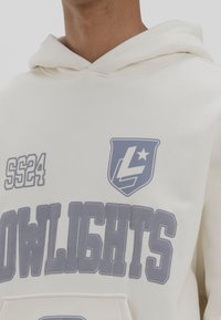 Zbliżenie osoby noszącej kremową bluzę z kapturem z napisem "SS24", logo tarczy z literą "L" i gwiazdą oraz dużymi blokowymi literami tworzącymi słowo "DOWLIGHTS".