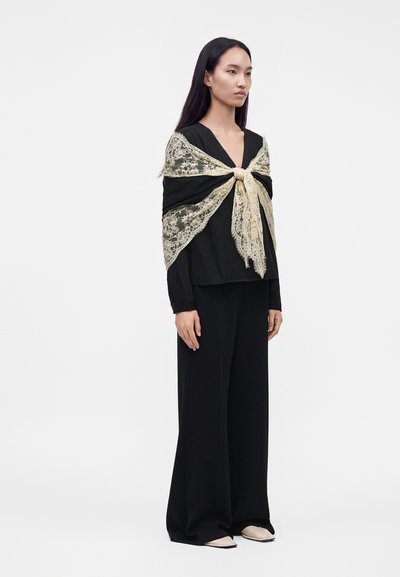 Femme portant un pantalon large noir et un haut avec un châle en dentelle crème transparente noué devant, debout devant un fond blanc uni.