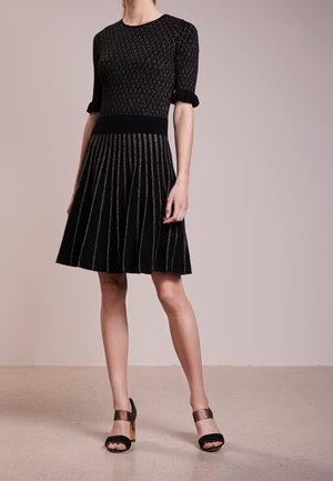 Mujer con un vestido negro hasta la rodilla, con la parte superior de patrón geométrico, falda plisada y tacones abiertos negros con tiras doradas en el tobillo.