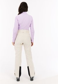 Donna che indossa pantaloni da neve beige con gaiters neri e una maglietta a maniche lunghe lilla, in piedi con la schiena rivolta verso un sfondo bianco.