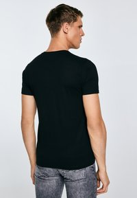 Vyras, stovintis nugara, dėvi paprastą juodą trumpomis rankovėmis marškinėlį ir šviesiai pilkus džinsus, prieš šviesiai pilką foną.