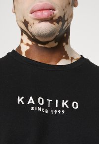 Sudadera negra con texto bordado "KAOTIKO" y "SINCE 1999" en blanco, con un cuello acanalado. Primer plano de la zona del pecho.