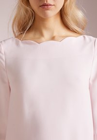 Ljusrosa blus med vågig halsringning, långa ärmar och mjuk textur. Tyget verkar mjukt och flödande, utan synliga mönster.