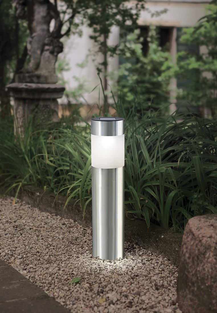 Lampa zewnętrzna ze stali nierdzewnej o cylindrycznym kształcie, z białym dyfuzorem szklanym i polerowanym wykończeniem, otoczona żwirem i zielenią.