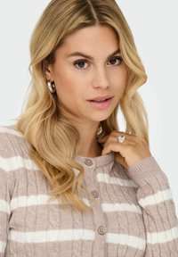 Lichtbruine kabelgebreide cardigan met witte strepen, met een ronde hals, knoopsluiting en ribbels aan de mouwen. Zilveren ring gedragen aan de hand.