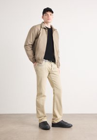 Hnědá bomber bunda přes černé tričko, béžové kalhoty a černé tenisky. Oblečení má jednoduchý design s volným střihem a bez vzorů.