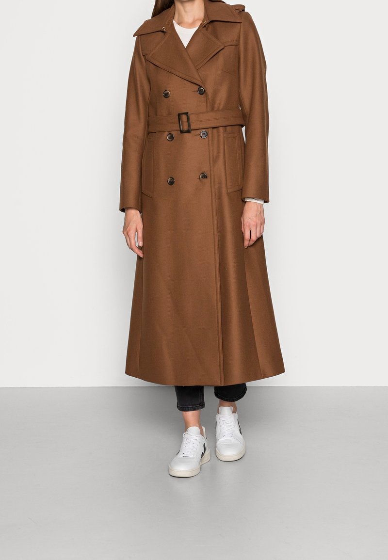Femme portant un long manteau marron croisé ceinturé à larges revers, un pantalon noir court et des baskets blanches, debout sur un sol gris.